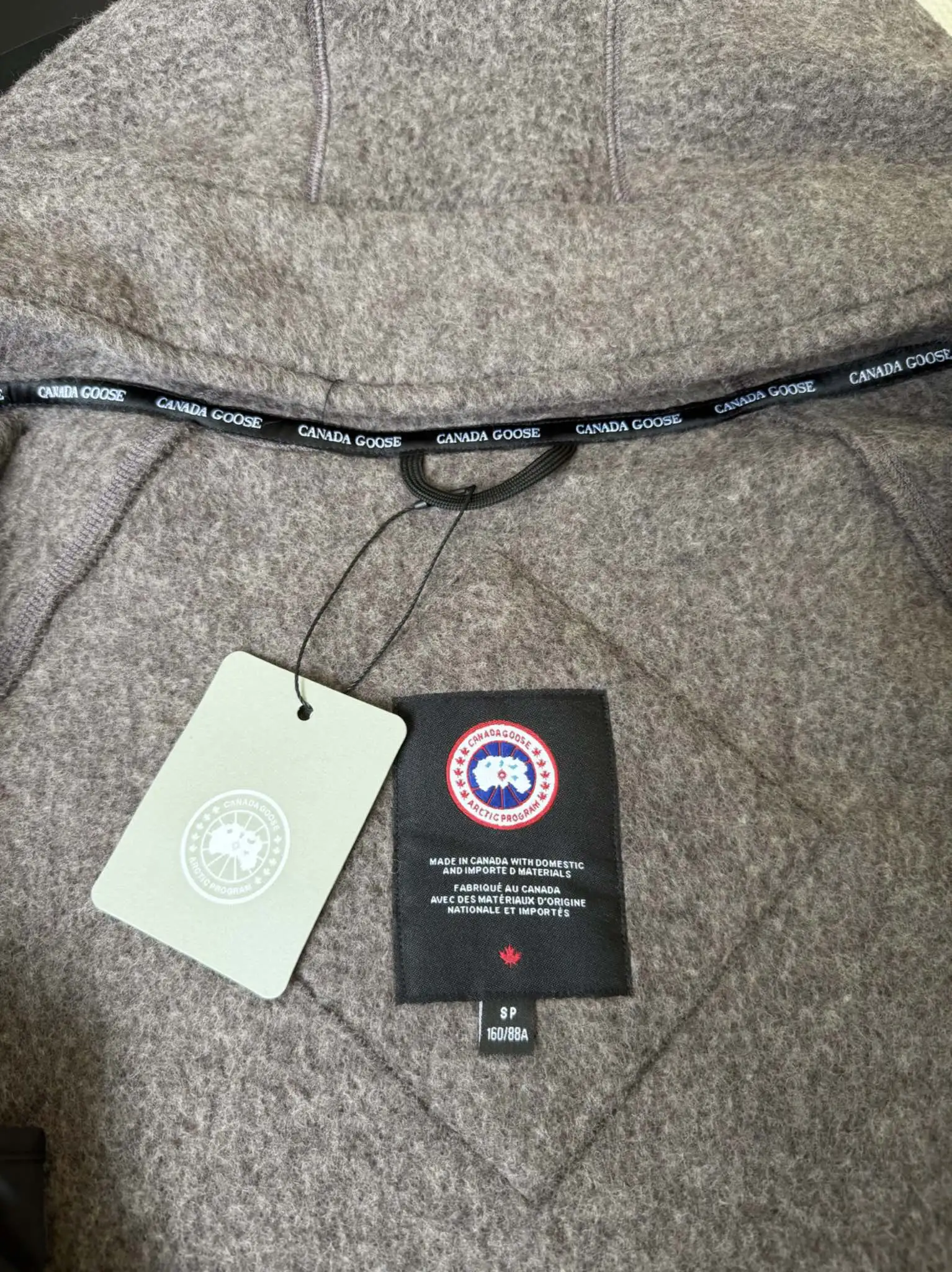 大鹅/Canada Goose抓绒羊毛饰章连帽夹克外套 面料选择: 以羊毛为主体，加入极少量氨纶和聚酯纤维混纺而成的羊毛抓绒面料。羊毛作为核心天然成分，提供无与伦比的温度调节、抑菌防臭和亲肤感。少量氨纶的喂入赋予了面料一定的四向弹性，日常活动时毫无束缚感。作为抓绒结构的基础，聚酯纤维增强了面料的耐久性、抗皱性和快干性，它构成了抓绒的蓬松感，并提供锁住空气等功能。 细节1: 衣袖和下摆采用2乘2罗纹收口，其强大的弹性，可以紧密贴合不同粗细的手腕和腰部，形成一个有效的&lsquo;密封&rsquo;,防止冷风倒灌。在多次拉伸和使用后仍能保持原有形状，不易松垮变形。且罗纹袖口能传递一种设计感，强化视觉美观度。 细节2: 兜帽设计与松紧扣的搭配提升穿戴时的保暖度与防风性能。 辅料:定制弹性抽绳松紧扣定制拉链吊坠定制臂章 尺码：S M L 颜色：黑 灰 咖 米 粉