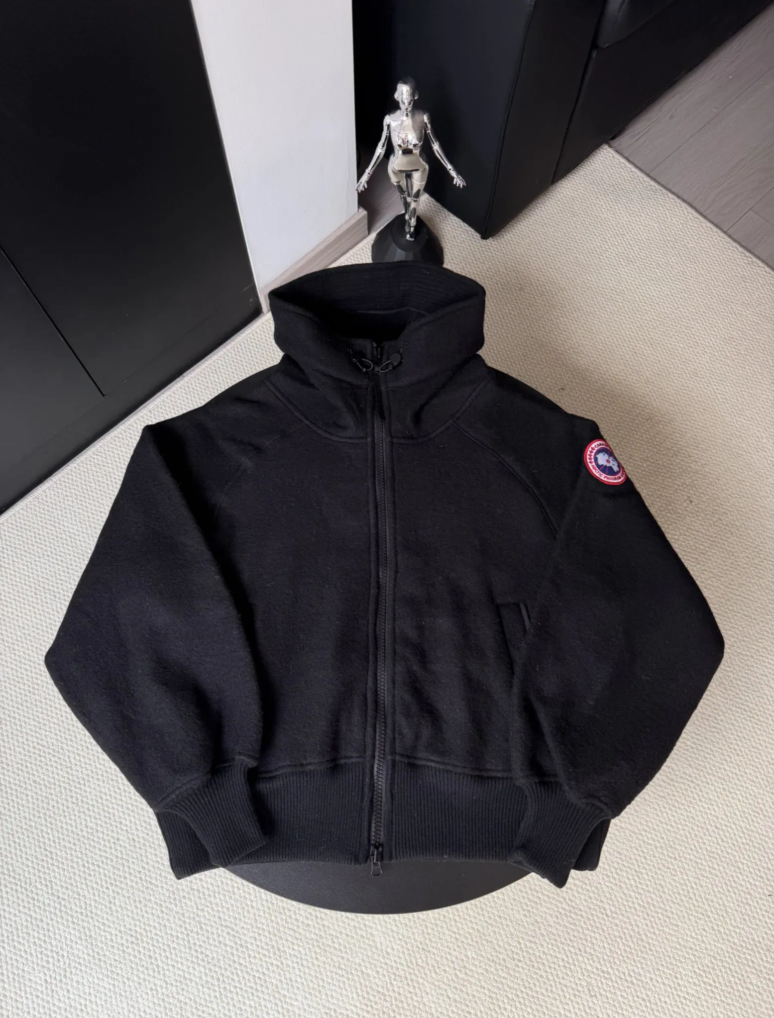 大鹅/Canada Goose抓绒羊毛饰章连帽夹克外套 面料选择: 以羊毛为主体，加入极少量氨纶和聚酯纤维混纺而成的羊毛抓绒面料。羊毛作为核心天然成分，提供无与伦比的温度调节、抑菌防臭和亲肤感。少量氨纶的喂入赋予了面料一定的四向弹性，日常活动时毫无束缚感。作为抓绒结构的基础，聚酯纤维增强了面料的耐久性、抗皱性和快干性，它构成了抓绒的蓬松感，并提供锁住空气等功能。 细节1: 衣袖和下摆采用2乘2罗纹收口，其强大的弹性，可以紧密贴合不同粗细的手腕和腰部，形成一个有效的&lsquo;密封&rsquo;,防止冷风倒灌。在多次拉伸和使用后仍能保持原有形状，不易松垮变形。且罗纹袖口能传递一种设计感，强化视觉美观度。 细节2: 兜帽设计与松紧扣的搭配提升穿戴时的保暖度与防风性能。 辅料:定制弹性抽绳松紧扣定制拉链吊坠定制臂章 尺码：S M L 颜色：黑 灰 咖 米 粉