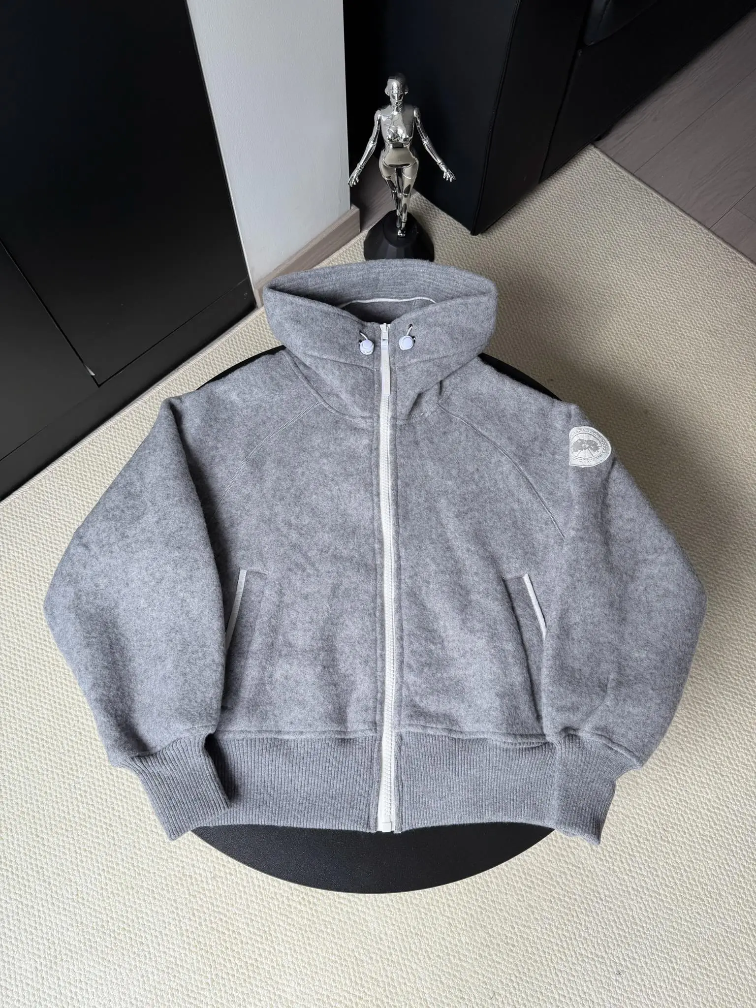 大鹅/Canada Goose抓绒羊毛饰章连帽夹克外套 面料选择: 以羊毛为主体，加入极少量氨纶和聚酯纤维混纺而成的羊毛抓绒面料。羊毛作为核心天然成分，提供无与伦比的温度调节、抑菌防臭和亲肤感。少量氨纶的喂入赋予了面料一定的四向弹性，日常活动时毫无束缚感。作为抓绒结构的基础，聚酯纤维增强了面料的耐久性、抗皱性和快干性，它构成了抓绒的蓬松感，并提供锁住空气等功能。 细节1: 衣袖和下摆采用2乘2罗纹收口，其强大的弹性，可以紧密贴合不同粗细的手腕和腰部，形成一个有效的&lsquo;密封&rsquo;,防止冷风倒灌。在多次拉伸和使用后仍能保持原有形状，不易松垮变形。且罗纹袖口能传递一种设计感，强化视觉美观度。 细节2: 兜帽设计与松紧扣的搭配提升穿戴时的保暖度与防风性能。 辅料:定制弹性抽绳松紧扣定制拉链吊坠定制臂章 尺码：S M L 颜色：黑 灰 咖 米 粉