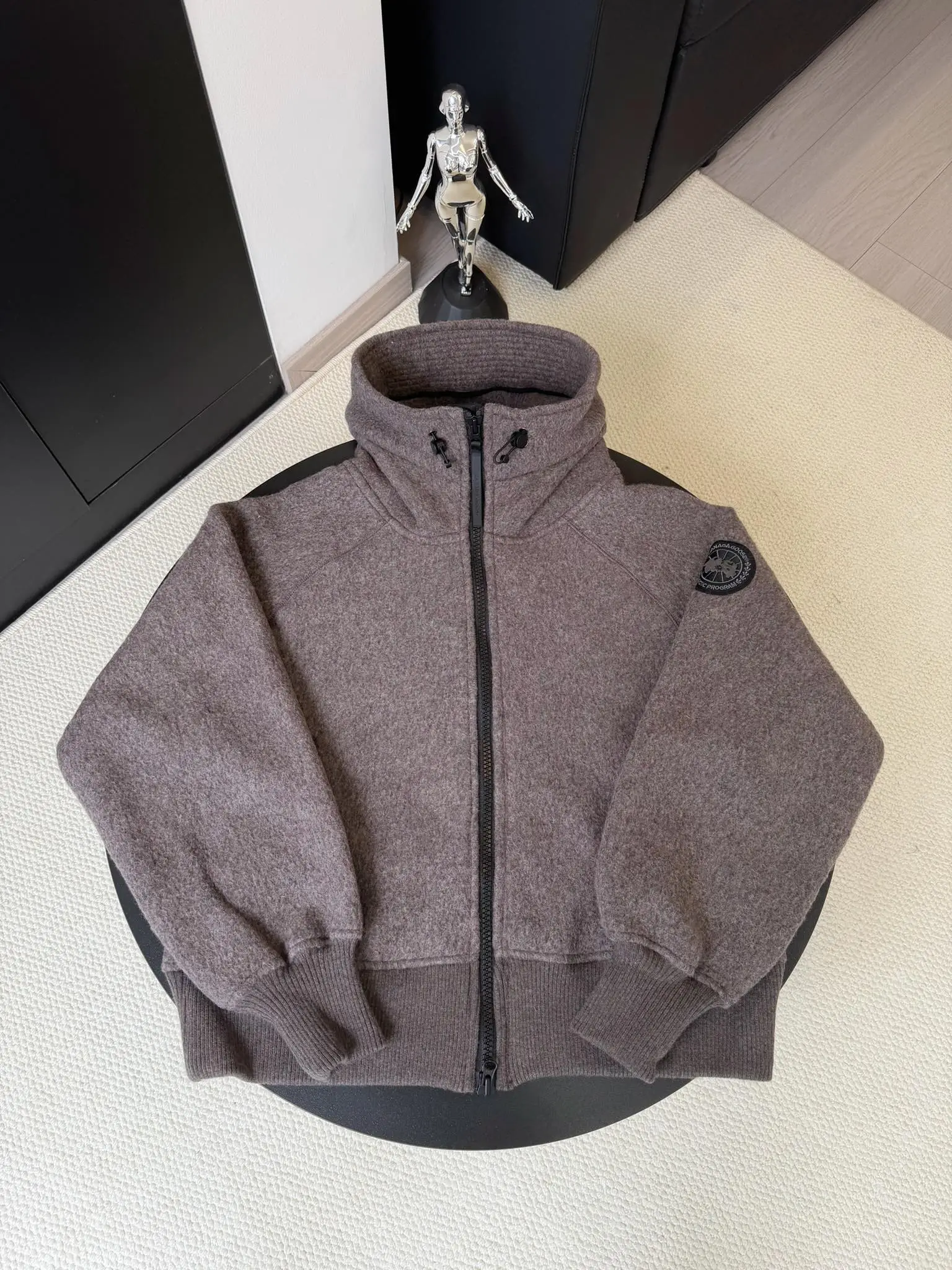 大鹅/Canada Goose抓绒羊毛饰章连帽夹克外套 面料选择: 以羊毛为主体，加入极少量氨纶和聚酯纤维混纺而成的羊毛抓绒面料。羊毛作为核心天然成分，提供无与伦比的温度调节、抑菌防臭和亲肤感。少量氨纶的喂入赋予了面料一定的四向弹性，日常活动时毫无束缚感。作为抓绒结构的基础，聚酯纤维增强了面料的耐久性、抗皱性和快干性，它构成了抓绒的蓬松感，并提供锁住空气等功能。 细节1: 衣袖和下摆采用2乘2罗纹收口，其强大的弹性，可以紧密贴合不同粗细的手腕和腰部，形成一个有效的&lsquo;密封&rsquo;,防止冷风倒灌。在多次拉伸和使用后仍能保持原有形状，不易松垮变形。且罗纹袖口能传递一种设计感，强化视觉美观度。 细节2: 兜帽设计与松紧扣的搭配提升穿戴时的保暖度与防风性能。 辅料:定制弹性抽绳松紧扣定制拉链吊坠定制臂章 尺码：S M L 颜色：黑 灰 咖 米 粉