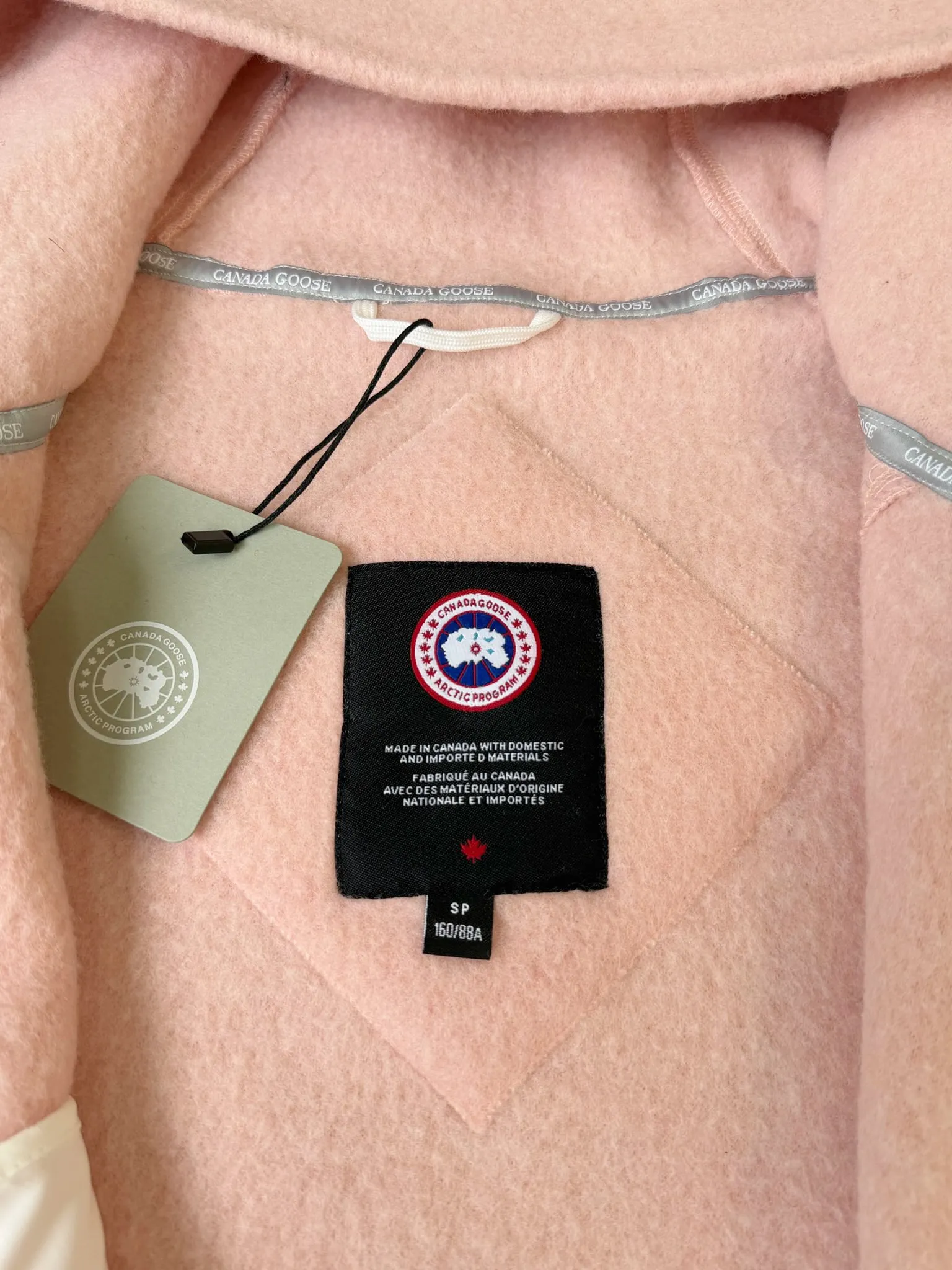 大鹅/Canada Goose抓绒羊毛饰章连帽夹克外套 面料选择: 以羊毛为主体，加入极少量氨纶和聚酯纤维混纺而成的羊毛抓绒面料。羊毛作为核心天然成分，提供无与伦比的温度调节、抑菌防臭和亲肤感。少量氨纶的喂入赋予了面料一定的四向弹性，日常活动时毫无束缚感。作为抓绒结构的基础，聚酯纤维增强了面料的耐久性、抗皱性和快干性，它构成了抓绒的蓬松感，并提供锁住空气等功能。 细节1: 衣袖和下摆采用2乘2罗纹收口，其强大的弹性，可以紧密贴合不同粗细的手腕和腰部，形成一个有效的&lsquo;密封&rsquo;,防止冷风倒灌。在多次拉伸和使用后仍能保持原有形状，不易松垮变形。且罗纹袖口能传递一种设计感，强化视觉美观度。 细节2: 兜帽设计与松紧扣的搭配提升穿戴时的保暖度与防风性能。 辅料:定制弹性抽绳松紧扣定制拉链吊坠定制臂章 尺码：S M L 颜色：黑 灰 咖 米 粉