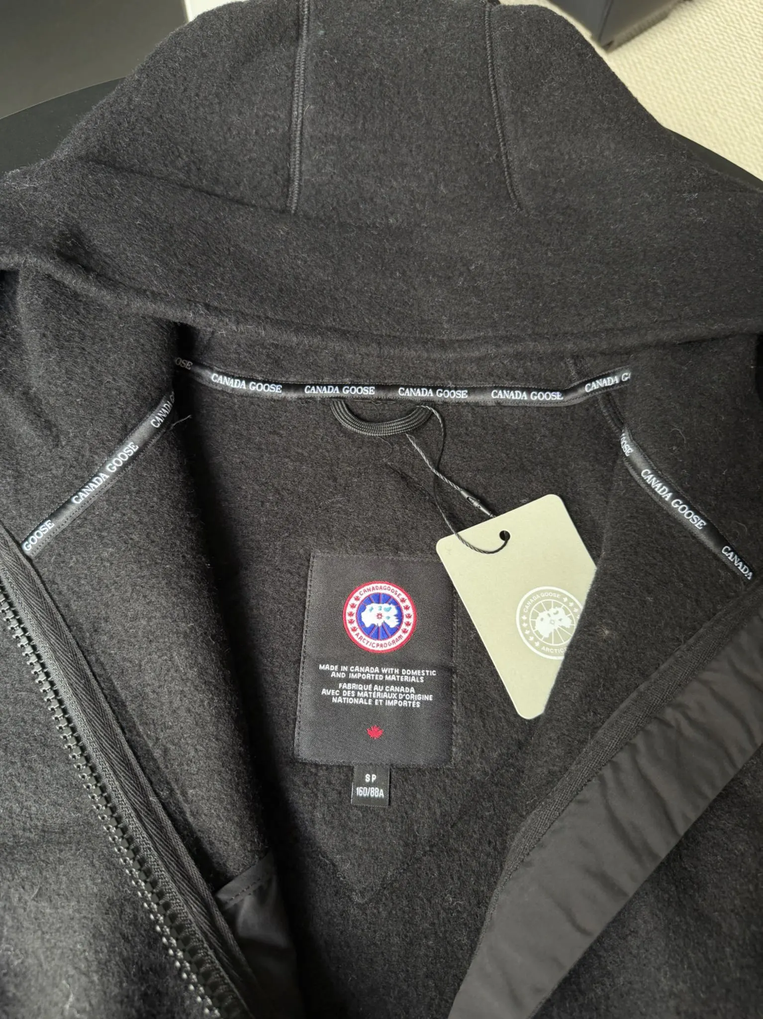 大鹅/Canada Goose抓绒羊毛饰章连帽夹克外套 面料选择: 以羊毛为主体，加入极少量氨纶和聚酯纤维混纺而成的羊毛抓绒面料。羊毛作为核心天然成分，提供无与伦比的温度调节、抑菌防臭和亲肤感。少量氨纶的喂入赋予了面料一定的四向弹性，日常活动时毫无束缚感。作为抓绒结构的基础，聚酯纤维增强了面料的耐久性、抗皱性和快干性，它构成了抓绒的蓬松感，并提供锁住空气等功能。 细节1: 衣袖和下摆采用2乘2罗纹收口，其强大的弹性，可以紧密贴合不同粗细的手腕和腰部，形成一个有效的&lsquo;密封&rsquo;,防止冷风倒灌。在多次拉伸和使用后仍能保持原有形状，不易松垮变形。且罗纹袖口能传递一种设计感，强化视觉美观度。 细节2: 兜帽设计与松紧扣的搭配提升穿戴时的保暖度与防风性能。 辅料:定制弹性抽绳松紧扣定制拉链吊坠定制臂章 尺码：S M L 颜色：黑 灰 咖 米 粉