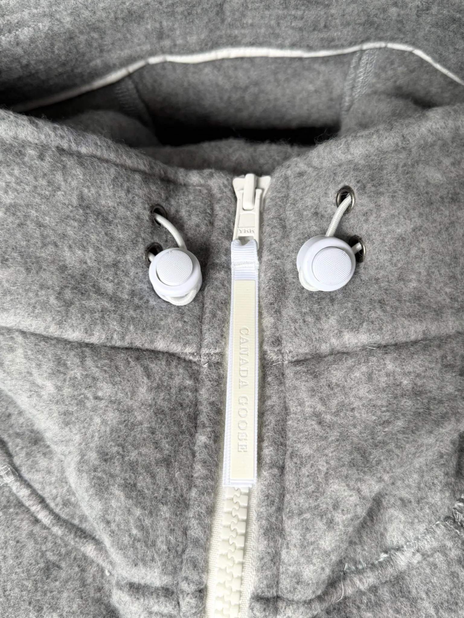 大鹅/Canada Goose抓绒羊毛饰章连帽夹克外套 面料选择: 以羊毛为主体，加入极少量氨纶和聚酯纤维混纺而成的羊毛抓绒面料。羊毛作为核心天然成分，提供无与伦比的温度调节、抑菌防臭和亲肤感。少量氨纶的喂入赋予了面料一定的四向弹性，日常活动时毫无束缚感。作为抓绒结构的基础，聚酯纤维增强了面料的耐久性、抗皱性和快干性，它构成了抓绒的蓬松感，并提供锁住空气等功能。 细节1: 衣袖和下摆采用2乘2罗纹收口，其强大的弹性，可以紧密贴合不同粗细的手腕和腰部，形成一个有效的&lsquo;密封&rsquo;,防止冷风倒灌。在多次拉伸和使用后仍能保持原有形状，不易松垮变形。且罗纹袖口能传递一种设计感，强化视觉美观度。 细节2: 兜帽设计与松紧扣的搭配提升穿戴时的保暖度与防风性能。 辅料:定制弹性抽绳松紧扣定制拉链吊坠定制臂章 尺码：S M L 颜色：黑 灰 咖 米 粉