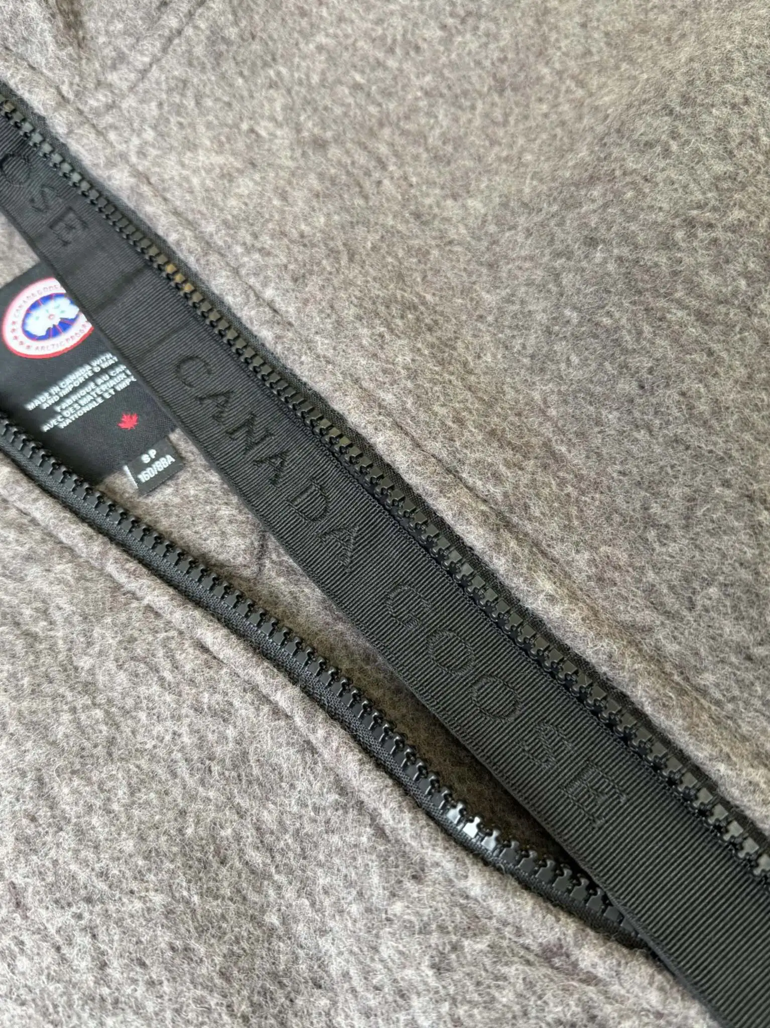 大鹅/Canada Goose抓绒羊毛饰章连帽夹克外套 面料选择: 以羊毛为主体，加入极少量氨纶和聚酯纤维混纺而成的羊毛抓绒面料。羊毛作为核心天然成分，提供无与伦比的温度调节、抑菌防臭和亲肤感。少量氨纶的喂入赋予了面料一定的四向弹性，日常活动时毫无束缚感。作为抓绒结构的基础，聚酯纤维增强了面料的耐久性、抗皱性和快干性，它构成了抓绒的蓬松感，并提供锁住空气等功能。 细节1: 衣袖和下摆采用2乘2罗纹收口，其强大的弹性，可以紧密贴合不同粗细的手腕和腰部，形成一个有效的&lsquo;密封&rsquo;,防止冷风倒灌。在多次拉伸和使用后仍能保持原有形状，不易松垮变形。且罗纹袖口能传递一种设计感，强化视觉美观度。 细节2: 兜帽设计与松紧扣的搭配提升穿戴时的保暖度与防风性能。 辅料:定制弹性抽绳松紧扣定制拉链吊坠定制臂章 尺码：S M L 颜色：黑 灰 咖 米 粉