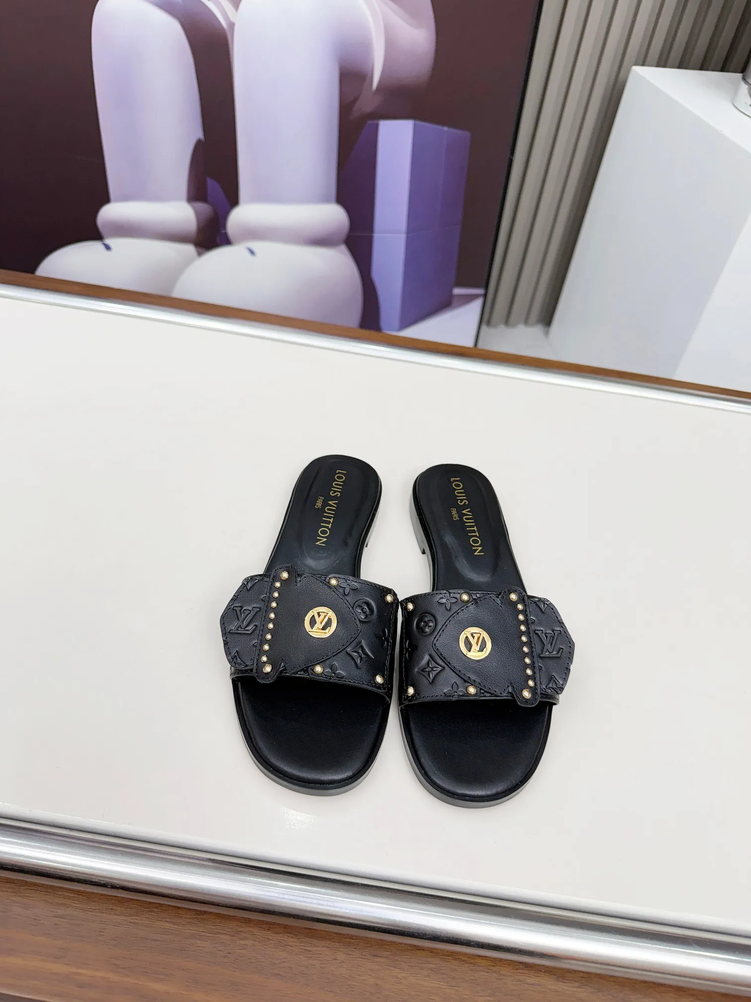 LV 1:1 premium Revival flat mules in black and white embossed Monogram leather with gold-tone studs and lock detail on display - xiashuluxe