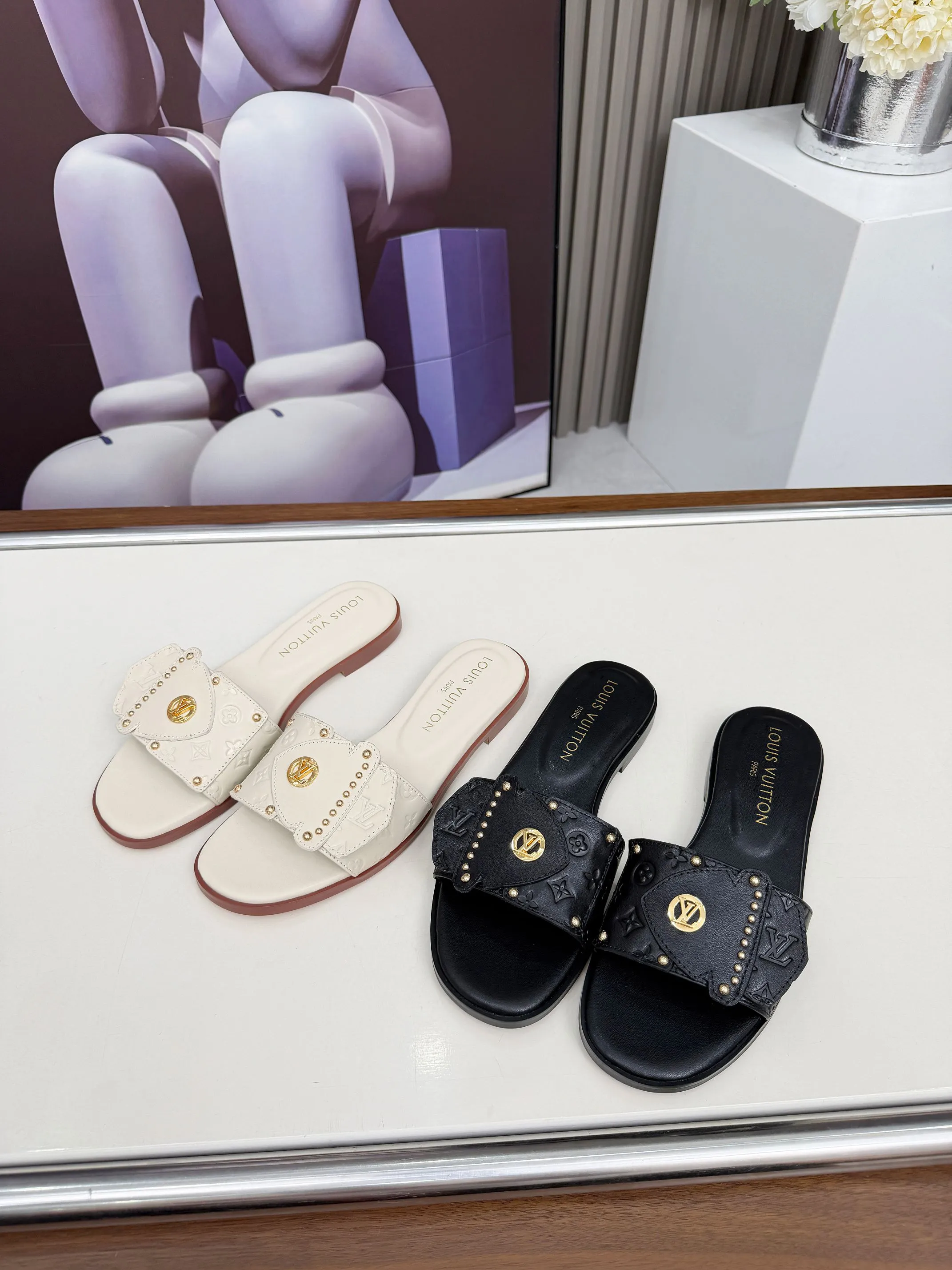 LV 1:1 premium Revival flat mules in black and white embossed Monogram leather with gold-tone studs and lock detail on display - xiashuluxe
