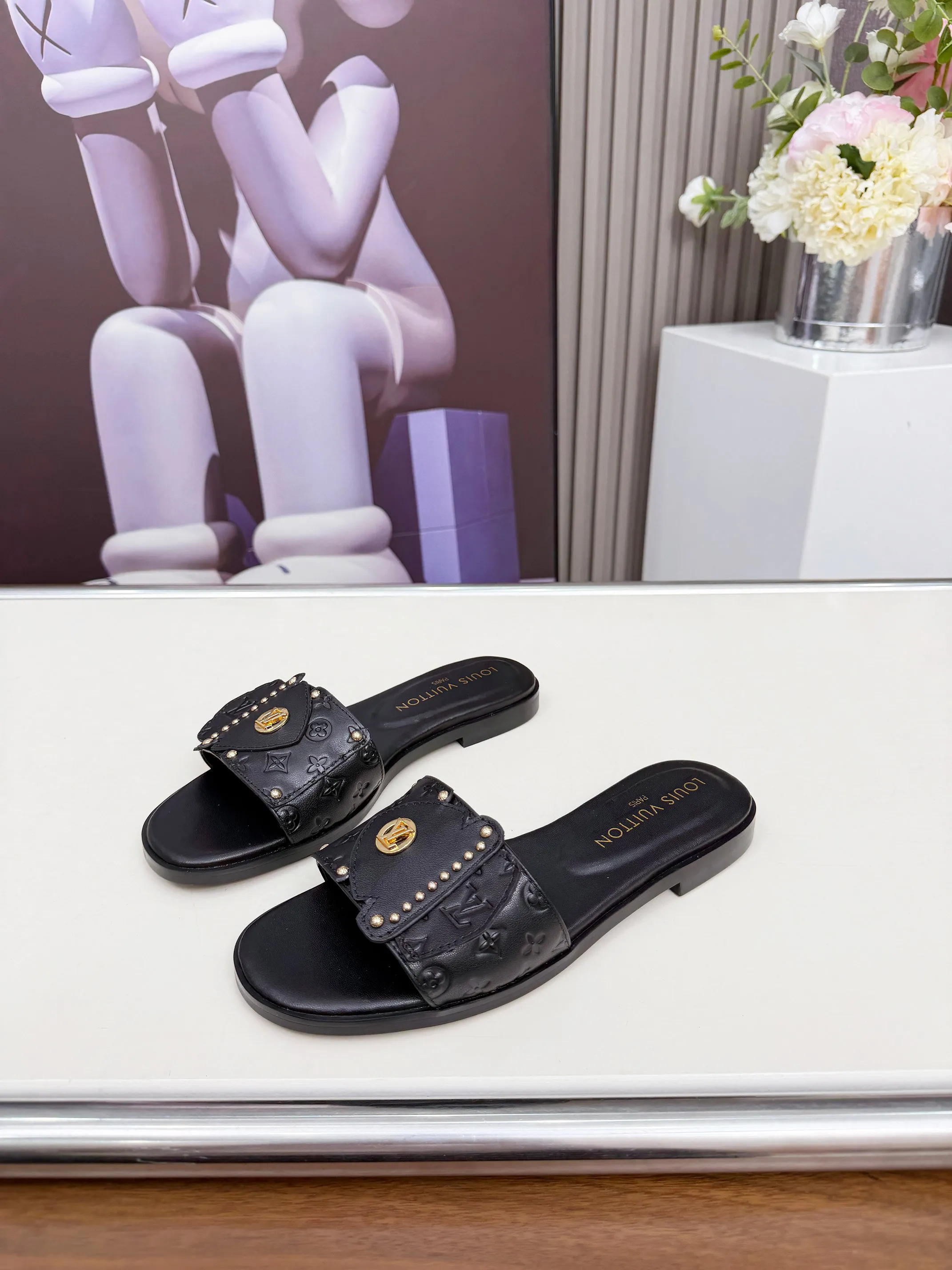 LV 1:1 premium Revival flat mules in black and white embossed Monogram leather with gold-tone studs and lock detail on display - xiashuluxe