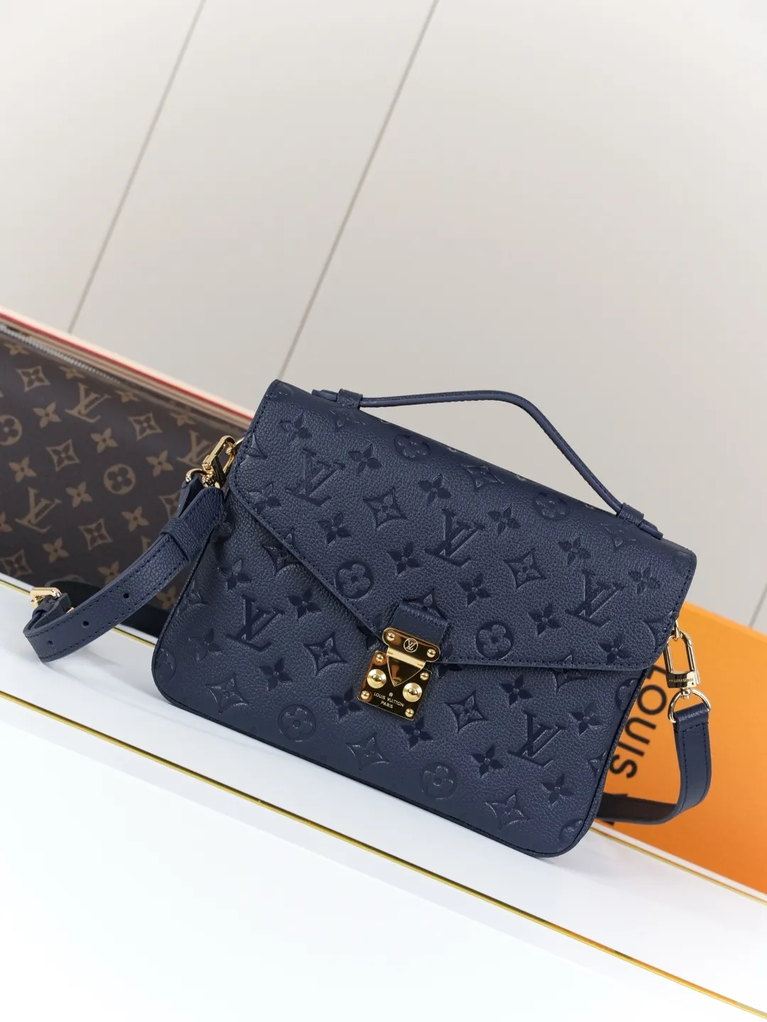 Front view of two Louis Vuitton Pochette Métis bags in Monogram Empreinte leather: one dark burgundy (Bordeaux) and one dark navy blue, placed on a white shelf with S-lock hardware, a partial Monogram bag and orange gift box background.