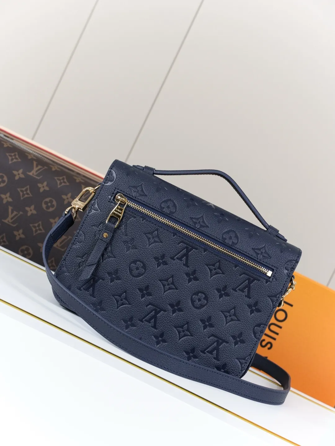 Front view of two Louis Vuitton Pochette M&eacute;tis bags in Monogram Empreinte leather: one dark burgundy (Bordeaux) and one dark navy blue, placed on a white shelf with S-lock hardware, a partial Monogram bag and orange gift box background.