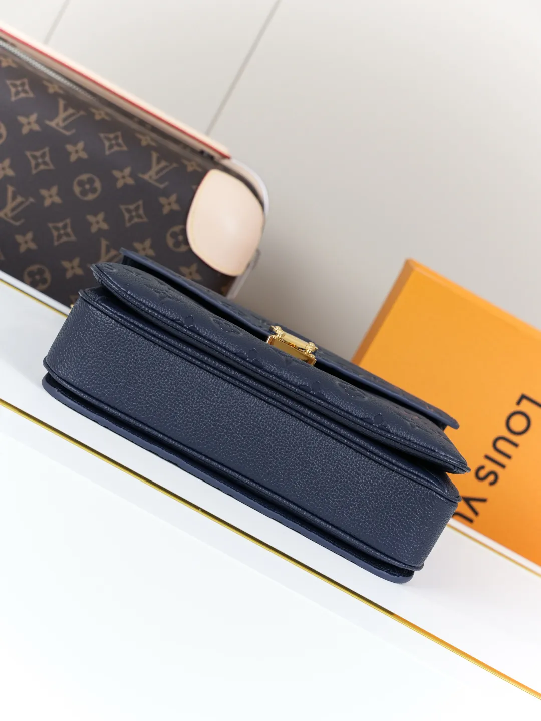 Front view of two Louis Vuitton Pochette M&eacute;tis bags in Monogram Empreinte leather: one dark burgundy (Bordeaux) and one dark navy blue, placed on a white shelf with S-lock hardware, a partial Monogram bag and orange gift box background.