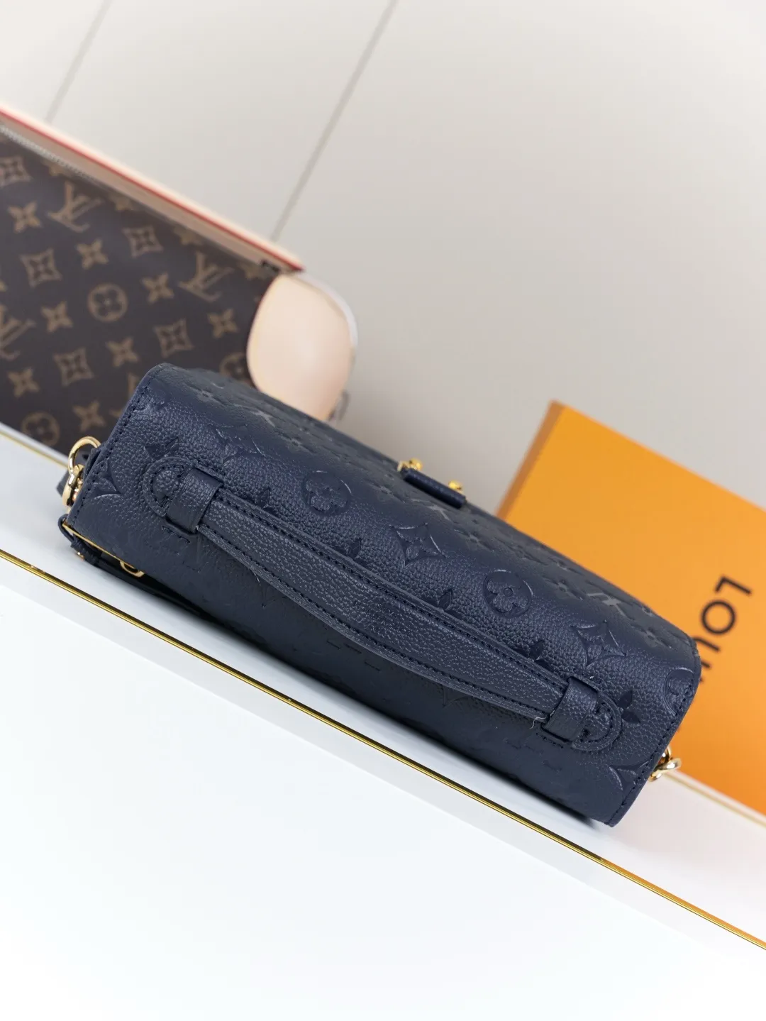 Front view of two Louis Vuitton Pochette Métis bags in Monogram Empreinte leather: one dark burgundy (Bordeaux) and one dark navy blue, placed on a white shelf with S-lock hardware, a partial Monogram bag and orange gift box background.