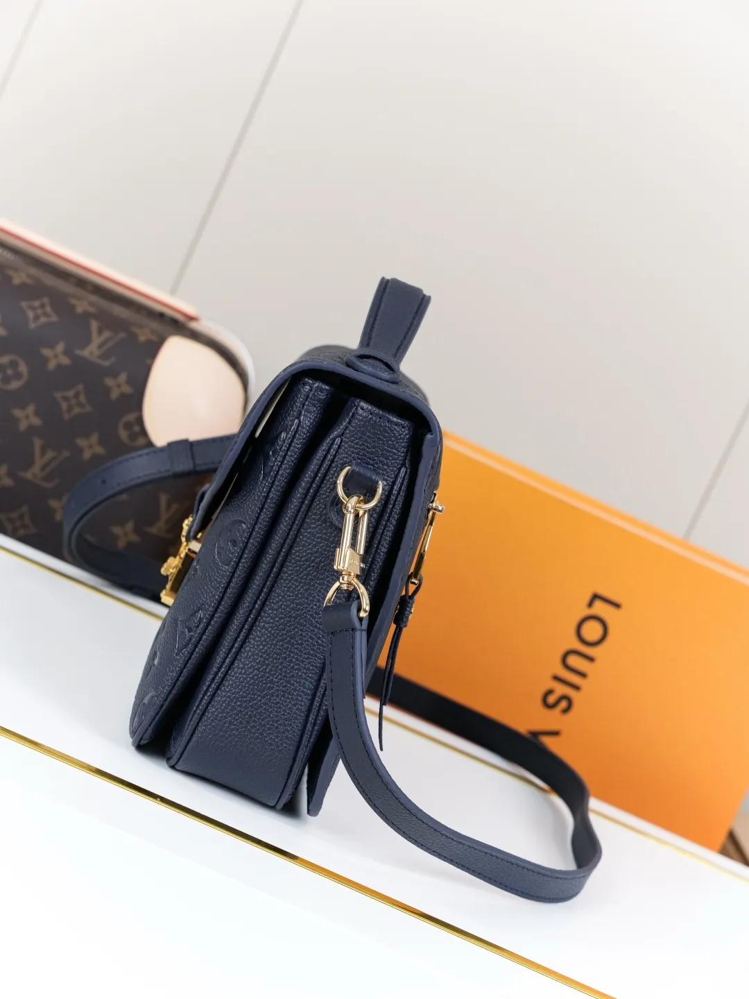 Front view of two Louis Vuitton Pochette M&eacute;tis bags in Monogram Empreinte leather: one dark burgundy (Bordeaux) and one dark navy blue, placed on a white shelf with S-lock hardware, a partial Monogram bag and orange gift box background.