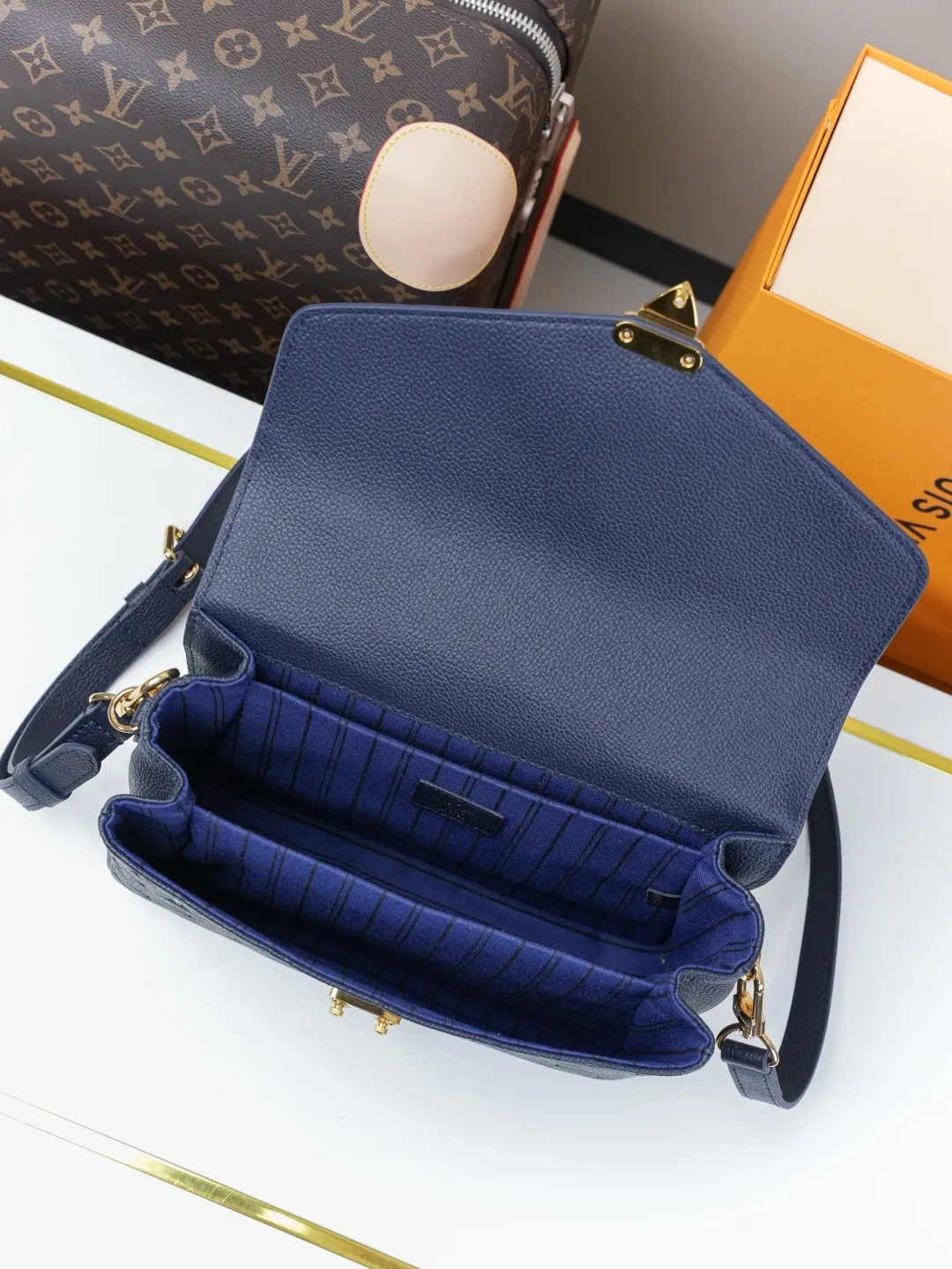 Front view of two Louis Vuitton Pochette M&eacute;tis bags in Monogram Empreinte leather: one dark burgundy (Bordeaux) and one dark navy blue, placed on a white shelf with S-lock hardware, a partial Monogram bag and orange gift box background.