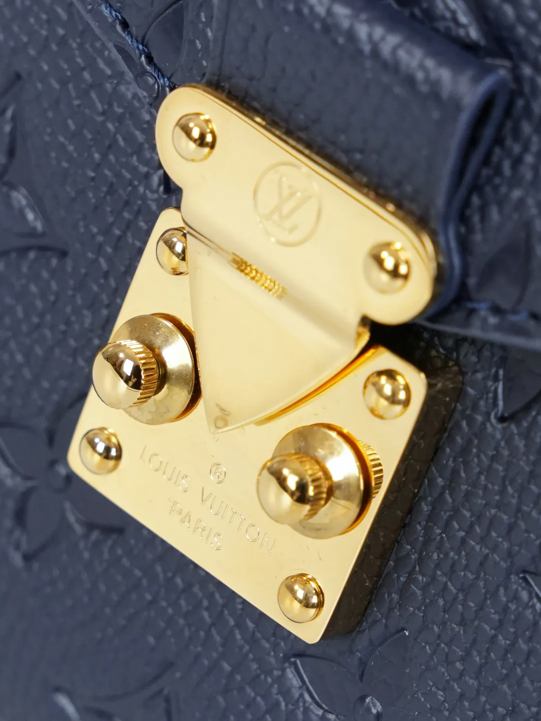 Front view of two Louis Vuitton Pochette M&eacute;tis bags in Monogram Empreinte leather: one dark burgundy (Bordeaux) and one dark navy blue, placed on a white shelf with S-lock hardware, a partial Monogram bag and orange gift box background.