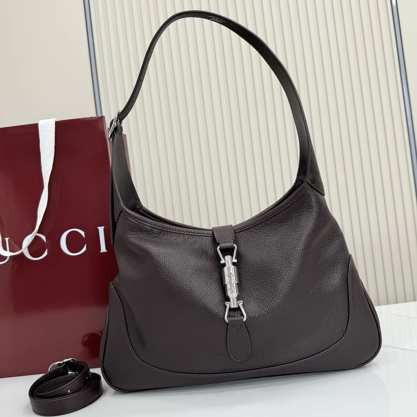 p750。【配全套包装】‼️（羊皮）2026春夏系列全新上市。这款Gucci Jackie 9161系列手袋呈现时髦风格，选用柔软粒面羊皮革，经手工悉心做旧处理，呈现出复古质感。手袋搭配便于使用的推入式锁扣设计，金色调金属配件更添品牌标志性五金细节的风格魅力。 型号：863136。 尺寸：31.5厘米（宽）x 22厘米（高）x 4.5厘米（深） 重量：约0.35千克 颜色：深棕色/羊皮。