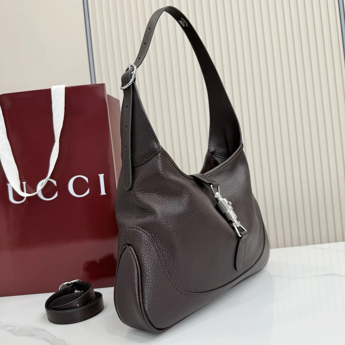 p750。【配全套包装】‼️（羊皮）2026春夏系列全新上市。这款Gucci Jackie 9161系列手袋呈现时髦风格，选用柔软粒面羊皮革，经手工悉心做旧处理，呈现出复古质感。手袋搭配便于使用的推入式锁扣设计，金色调金属配件更添品牌标志性五金细节的风格魅力。 型号：863136。 尺寸：31.5厘米（宽）x 22厘米（高）x 4.5厘米（深） 重量：约0.35千克 颜色：深棕色/羊皮。